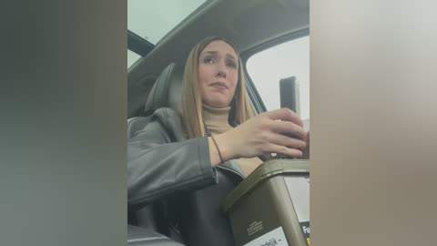 Media: Video of a woman with light skin, straight blonde hair, wearing a black leather jacket and a beige turtleneck, driving a car with beige interior.