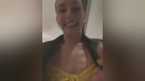 Media: Video of a smiling woman with dark hair, wearing a yellow lace bra and a green jacket, partially obscured by out-of-focus, blurry objects in the foreground.