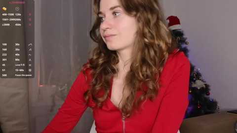 Media: Video of a young woman with long, wavy brown hair, wearing a red zip-up hoodie, looking contemplative, in a softly lit room with a Christmas tree in the background.