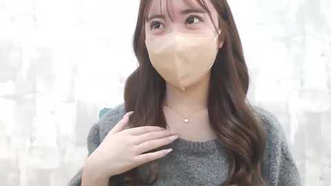 Media: A video of a young Asian woman with long brown hair, wearing a gray sweater and a beige face mask, standing against a blurred, light-colored background.