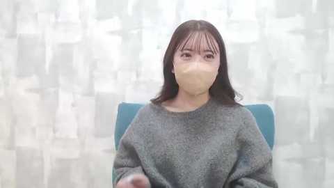 Media: Video of an Asian woman with straight black hair, wearing a gray sweater and beige surgical mask, seated in a teal chair against a white and gray patterned backdrop.