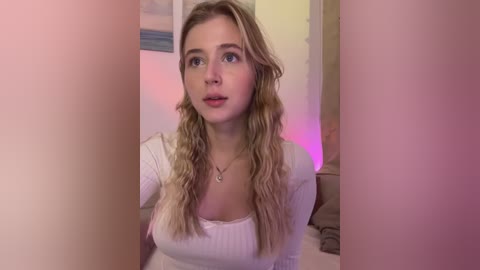 Media: A video of a young Caucasian woman with long, wavy blonde hair, fair skin, and blue eyes, wearing a white ribbed top, sitting in a dimly lit room with pink lighting and a framed painting on the wall.