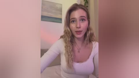 Media: Video of a young Caucasian woman with wavy blonde hair, wearing a white ribbed long-sleeve top, sitting on a beige couch in a softly lit room with a framed painting in the background.