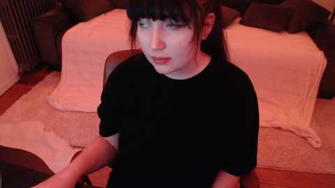 Media: A video of a young Asian woman with pale skin, dark hair, and a black t-shirt, sitting on a chair in a dimly lit bedroom with a bed covered in white sheets.