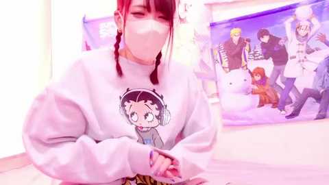 Media: Video of a young woman with red and black braided hair, wearing a white hoodie with a cartoon character and a face mask, in a room with anime posters.