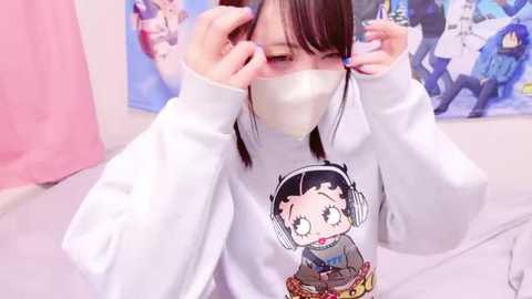 Media: Video of an Asian woman with straight black hair, wearing a white sweatshirt featuring a cartoon character, adjusting her face mask in a room with pastel walls and anime posters.