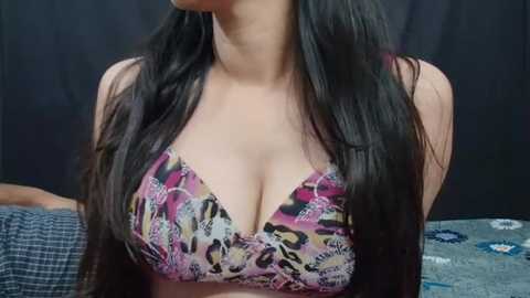 Media: Video of a woman with long, straight black hair, wearing a colorful floral-patterned bra. She has light skin and is indoors against a dark backdrop.