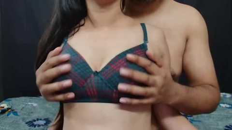 Media: A video captures an Asian woman with long black hair, wearing a green and purple plaid bra, being held by a man's hands from behind. The background is a dark room with a bed and a black curtain.