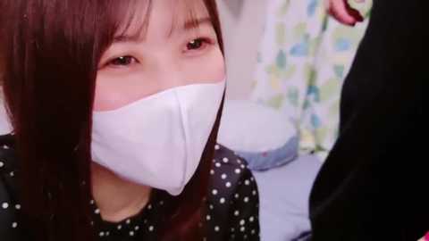 Media: Video of an Asian woman with straight brown hair and light skin, wearing a white surgical mask and black polka-dotted gown, seated in a medical room with colorful wall art.