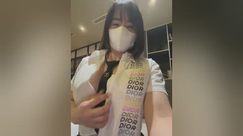 Media: Video of an Asian woman with short black hair, wearing a white mask, a black top, and a Dior robe with \"Dior\" text. She is indoors, standing in a modern, well-lit room with white walls and recessed lighting.