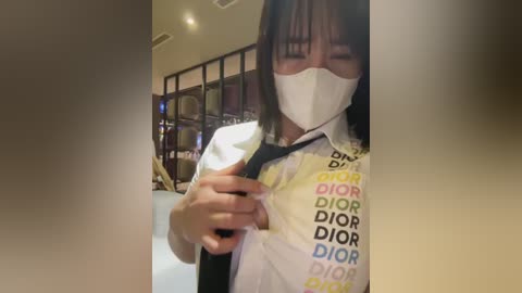 Media: Video of an Asian woman in a Dior shirt, wearing a surgical mask, adjusting a black tie, indoors.