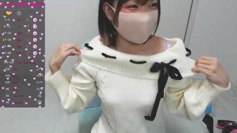 Media: Video of an East Asian woman with short brown hair, wearing a white knit sweater with black bows and a face mask, in a sterile white room with a blue chair, captured from a low angle.