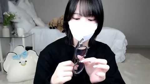 Media: Video of a young Asian woman with straight black hair, wearing a white mask, adjusting glasses, in a cozy bedroom with a teddy bear and flowers on a glass table.