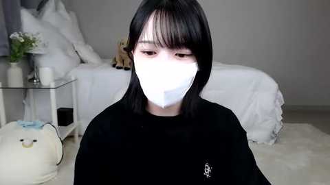 Media: Video of an Asian woman with straight black hair, wearing a white face mask and black top, in a minimalist bedroom with a white bed, beige carpet, and a small white nightstand.