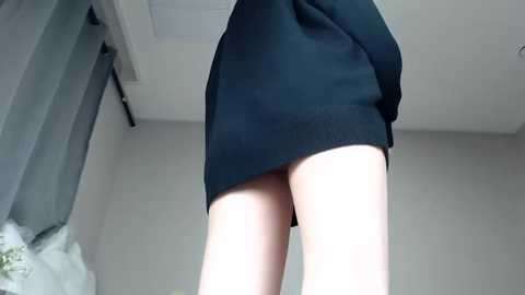 Media: Video of a woman's bare legs, wearing a short, black dress, standing indoors against a plain, light-colored wall.