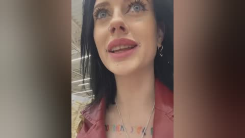 Media: A close-up video of a smiling young woman with pale skin, blue eyes, and black hair, wearing a red jacket and a colorful scarf. She has a small nose stud and a thin silver necklace. The background is blurry.