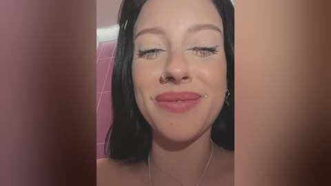 Media: Video of a smiling young woman with long black hair, wearing a nose ring and subtle makeup, in a bathroom with pink tiles.