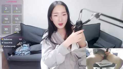 Media: A video captures an Asian woman with long black hair, light skin, and a slender physique, sitting on a couch. She is holding a black wig, looking relaxed. A TV screen in the background shows her live stream.