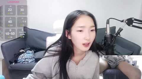 Media: Video of a young East Asian woman with long black hair, wearing a gray shirt, sitting in a modern living room with a gray sofa, a white wall calendar, and a microphone.