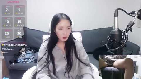 Media: Video of a young Asian woman with long black hair, wearing a grey sweatshirt, sitting on a bed. A microphone is on a stand beside her, and a blue and white plaid blanket is on the bed.