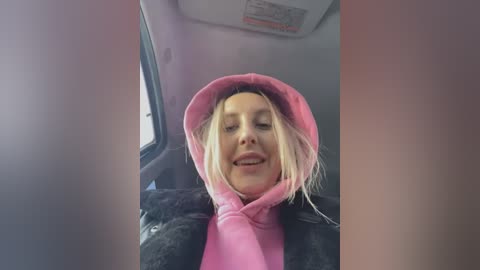 Media: Video of a smiling woman with long blonde hair, wearing a pink hoodie inside a car. The interior is dimly lit, with a window visible to the left.