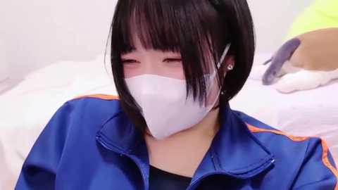 Media: Video of an Asian woman with short black hair, wearing a white surgical mask, blue and orange jacket, and lying on a bed with white sheets and a yellow pillow.