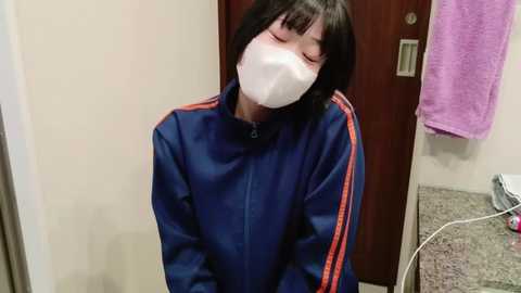 Media: Video of an Asian woman with short black hair, wearing a blue tracksuit with red stripes, white face mask, and closed eyes, standing in a bathroom with a purple towel hanging and a white cord on the floor.