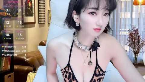 Media: A video of a young Asian woman with short black hair, wearing a leopard-print halter dress, sitting on a white couch in a cozy living room with framed art and a potted plant.