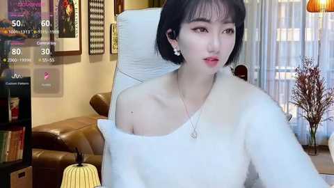 Media: Video of an East Asian woman with short black hair, wearing a white off-shoulder sweater, sitting on a brown leather couch. Background includes framed artwork, a TV screen, and a plant.