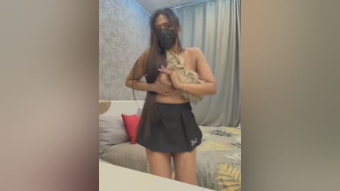 Media: Video of a young woman with long dark hair, wearing a black face mask, holding a small dog, standing in a bedroom with gray walls, white bed, and yellow pillow.