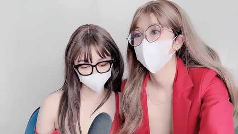 Media: Video of two women wearing white face masks, black-framed glasses, and red blazers, with long, straight, light brown hair. They are seated against a plain, light gray background.