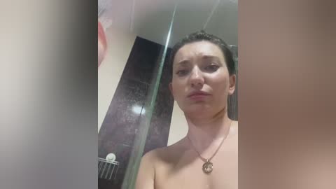 Media: Video of a woman with fair skin, wet hair, and bare shoulders, wearing a gold necklace, standing in a shower with black tiled walls.