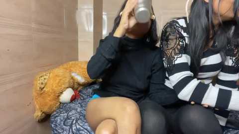 Media: Video of a young woman in a black turtleneck, drinking from a silver cup, with a stuffed animal and a patterned blanket beside her, in a cozy, beige-walled room.