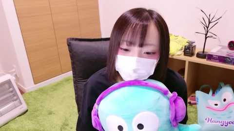 Media: Video of an East Asian woman wearing a face mask, seated on a black chair, holding a plush toy. Background includes a green carpet, wooden furniture, and a bookshelf.