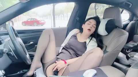 Media: A video of an Asian woman, in her 20s, reclining in a car, wearing a tight, black dress, white jacket, and sheer stockings, with her legs spread, revealing her shaved vulva.