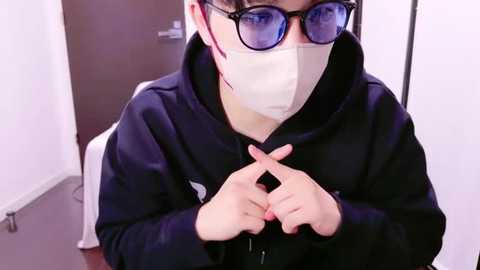 Media: Video of an Asian person wearing glasses, a face mask, and a black hoodie, seated indoors, with a white wall and door in the background.