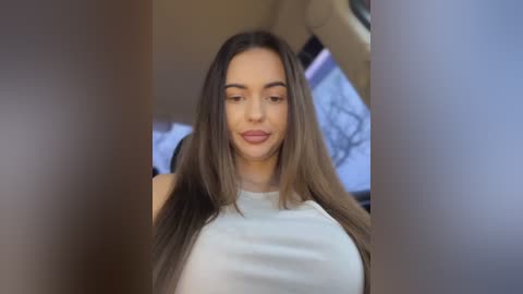 Media: Video of a young woman with long, straight brown hair, fair skin, and a slim physique, wearing a white sleeveless top. She has a serene expression and is seated in the passenger seat of a car.