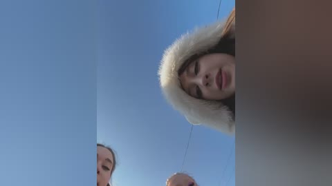 Media: A video of three people looking upwards at a clear blue sky, one woman in a white fur-lined hooded coat and another woman and man in the background.