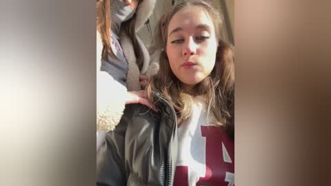 Media: Video: A young woman with light brown hair, wearing a grey sweatshirt and black jacket, sits indoors, looking contemplative.