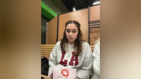 Media: Video of a young woman with long brown hair, wearing a light grey hoodie with \"LA\" in red, sitting in a locker room. She holds a pink bag with a red star.