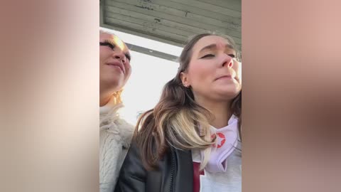 Media: Video of two young women, one in a grey coat, the other in a white blouse and pink scarf, standing under a wooden awning, smiling and looking at each other.