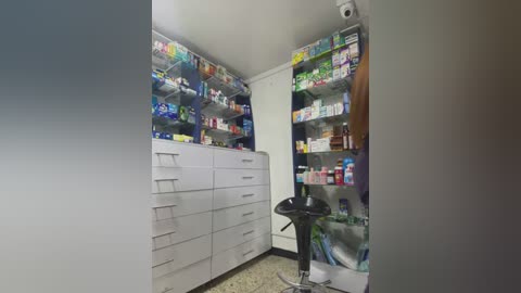 Media: Video of a compact, cluttered room with white cabinets filled with various medicines, a small black stool, and a speckled floor.