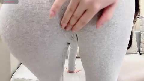 Media: A video of a woman's lower body, showing her hand spreading her grey, tight-fitting leggings, revealing a dildo inserted into her vagina. Background includes a white couch and light-colored walls.