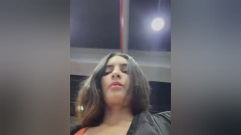 Media: Video of a Latina woman with medium skin tone, shoulder-length brown hair, and full lips, wearing a black top, standing indoors near a window with a red frame and a bright light source.