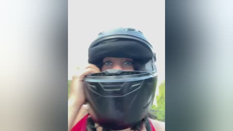 Media: Video of a person in a full-face black motorcycle helmet, partially obscuring their face. They have light skin and are wearing a red top. The background is blurred, with hints of greenery.