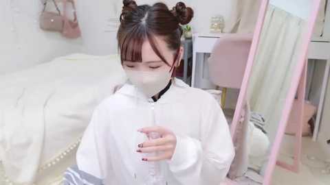 Media: Video of an East Asian woman with dark hair in buns, wearing a white surgical mask, white lab coat, and gloves, sitting on a beige carpet in a minimalist room with white walls, a bed, and a desk.