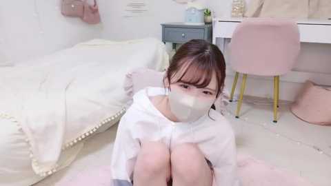 Media: A video of an Asian woman with light skin and brown hair, wearing a white mask and a loose white top, sitting on a soft pink rug in a pastel-colored bedroom with white furniture.