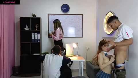 Media: Video of a white man in a white t-shirt, shirtless, performing oral sex on a topless woman with red hair, while another man in glasses watches. Background includes a white wall with a round clock, shelves, and a desk.