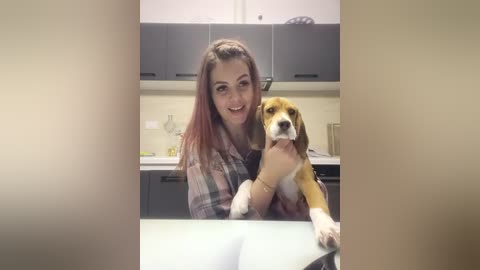 Media: Video of a smiling woman with long, reddish-brown hair, wearing a plaid shirt, holding a brown and white beagle in a modern kitchen with gray cabinets and white countertops.