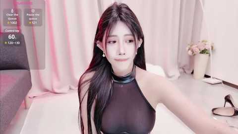 Media: A video of an East Asian woman with long black hair, wearing a sheer black halter top, sitting on a white couch in a minimalist room.